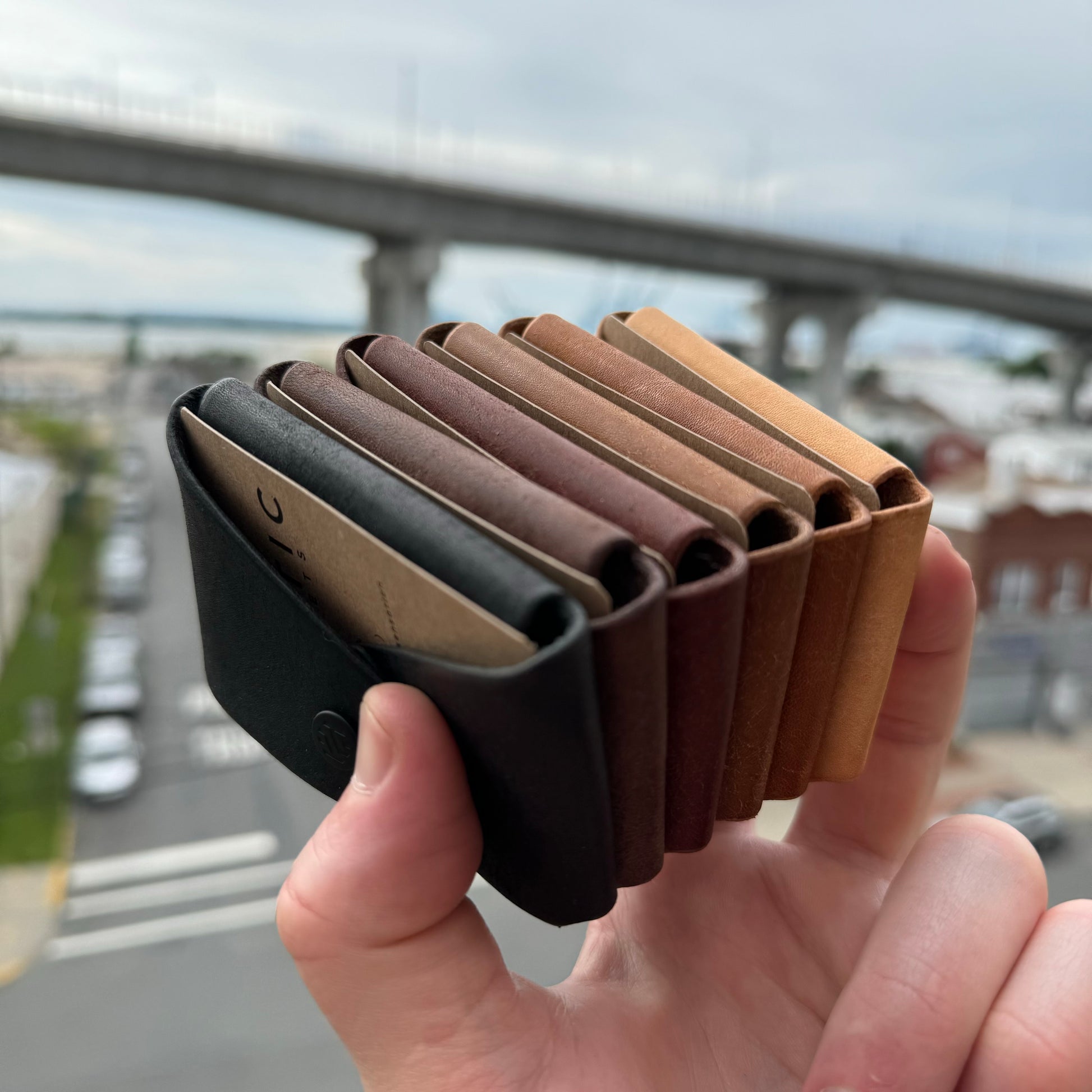 A hand holding multiple minimalist leather wallets in different colors, with an urban backdrop.